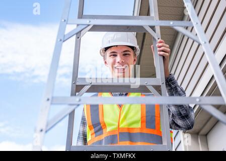 Giovane architetto maschio in piedi su una scaletta e vestito di ingranaggio di sicurezza. Foto Stock