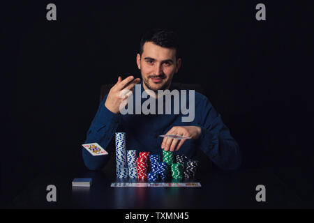 Fiducioso Ragazzo giocatore di poker seduti al tavolo del casinò di gettare le carte da gioco su sfondo nero. Il gioco d'azzardo vincitore torneo concetto di successo. Foto Stock