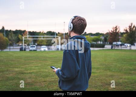 Adolescente ascoltare musica con le cuffie. Egli è in piedi al centro di un campo di calcio vicino a una scuola. Foto Stock