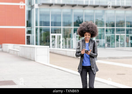 Giovane donna con capelli afro in città, utilizzando il touchscreen dello smartphone Foto Stock