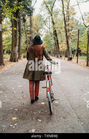 Giovane donna con lunghi capelli rossi spingendo in bicicletta in autunno park, vista posteriore a lunghezza piena, Firenze, Toscana, Italia Foto Stock