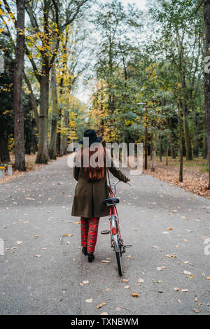 Giovane donna con lunghi capelli rossi spingendo in bicicletta in viale alberato autunno park, vista posteriore, Firenze, Toscana, Italia Foto Stock