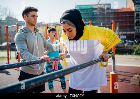 Classe Calisthenics presso palestra all'aperto, maschio formatore incoraggiando giovane donna sulle barre parallele Foto Stock