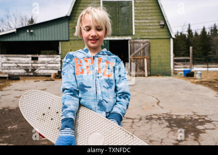 Ragazzo biondo che trasportano skateboard in cortile, ritratto Foto Stock