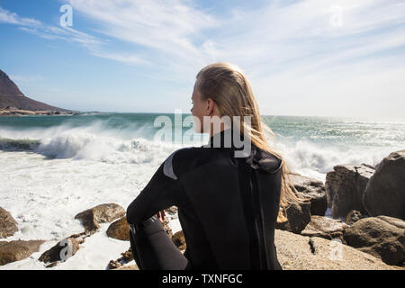 Giovani femmine surfer con lunghi capelli biondi seduto sulla spiaggia rock, vista posteriore, Cape Town, Western Cape, Sud Africa Foto Stock