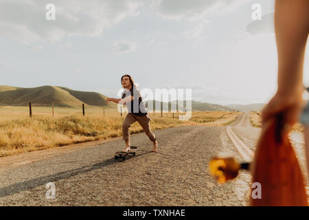 Giovane maschio a piedi nudi guidatore di skateboard skateboard su strada rurale, fidanzata a guardare, Exeter, CALIFORNIA, STATI UNITI D'AMERICA Foto Stock