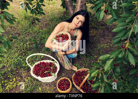 Giovane e bella donna che indossa abiti colorati sotto la struttura cherriy con due cesti di vimini e due secchi pieni di frutta messa a fuoco selettiva Foto Stock