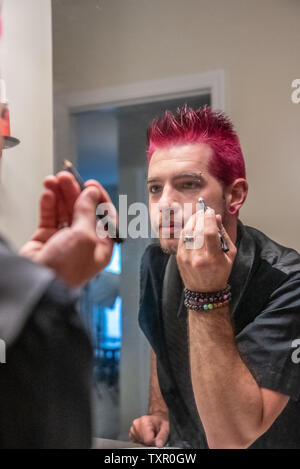 Diversi caucasici uomo maschio con dentato per capelli rosa applicazione eyeliner specchio dello specchio di riflessione. Indossa abiti neri con punk rocker l alternativa Foto Stock