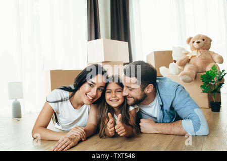 Bambino felice con la famiglia che mostra i pollici fino nella loro nuova casa. È meraviglioso il passaggio a un nuovo appartamento Foto Stock