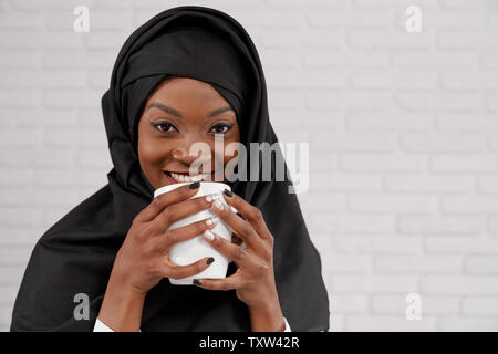 Bella e affascinante donna musulmana holding tazza bianca, con sorriso perfetto guardando la fotocamera. Piuttosto, giovane ed elegante ragazza africana di nero e di bianco manicure vestita di nero hijab. Foto Stock