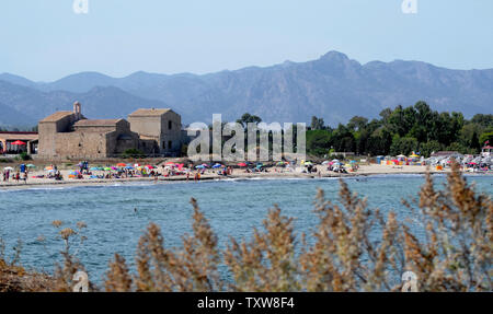 Sardegna, Italia, Agosto 2017, la baia di Nora con i turisti sulla spiaggia, il borgo medievale di Sant'Efisio chiesa vicino la riva e le montagne sullo sfondo Foto Stock