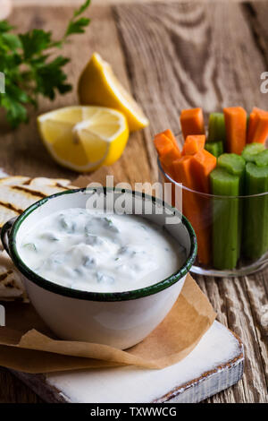 Yogurt dip con il prezzemolo e il succo di limone servita su tavola in legno rustico con tortilla chips e verdure fresche di carota e sedano, vegetar sana Foto Stock