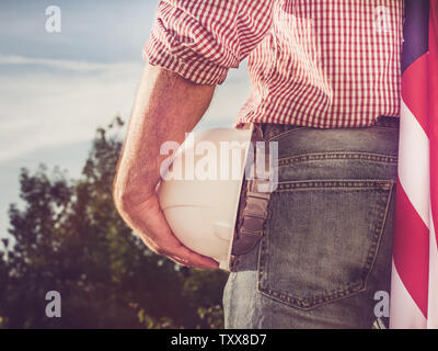 Bello ingegnere, azienda hardhat bianco e una bandiera americana nel parco contro lo sfondo di alberi verdi e il sole di setting, esaminando il di Foto Stock