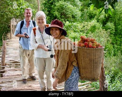 Una coppia europea sorridente posa per foto sul ponte in campagna con amichevole donna vietnamita che porta cesto di frutta di rambutan sulla schiena Foto Stock