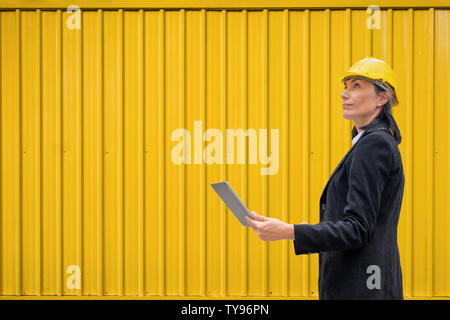 Leader della donna ingegnere con casco stand portatile tenuta sul contenitore giallo sullo sfondo Foto Stock