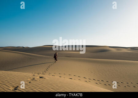 Suggestiva fiducioso giovane donna che cammina sul suo percorso sul deserto di sabbia con abito rosso in middlw di dune su caldo giorno d'estate con cielo blu chiaro Foto Stock