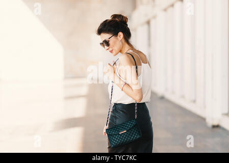 Bella sorridente giovane donna in occhiali da sole che indossa la frizione in pelle e mantello bianco facile camicetta e guardando lontano sopra la spalla mentre si cammina superarvi Foto Stock