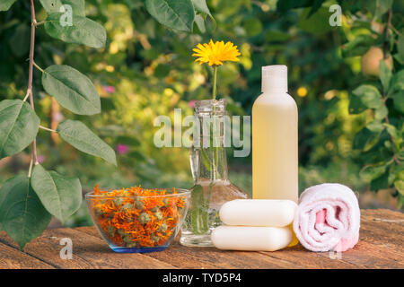 Prodotti per il benessere e gli accessori. Asciugamani, sapone e la bottiglia di plastica con estratto di calendula e di calendula fiori in un pallone di vetro su tavole di legno. Foto Stock