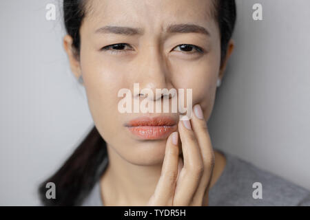 La donna mise la sua mano sulla guancia a causa di mal di denti. Donna asiatica soffre di mal di denti. Giovane donna con di mal di denti su sfondo grigio. Foto Stock