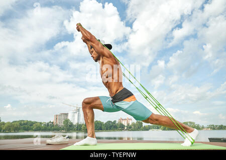 Sport professionali uomo tirando verso l'alto espansore e affondo facendo esercizio da gambe. Maschio nel breve lavoro fuori in mattinata vicino al lago, stretching sul materassino yoga. Foto Stock