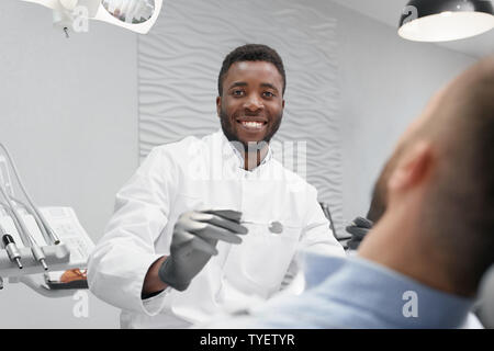 Messa a fuoco selettiva di allegro dentista maschio nel processo di indurimento di denti in studio dentistico. Medico professionale guardando la fotocamera e ridere mentre il paziente sdraiato sulla poltrona del dentista. Concetto di stomatologia. Foto Stock