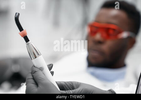 Messa a fuoco selettiva del dispositivo a raggi ultravioletti in mani femminili di dentista in clinica. Medico denti di indurimento, facendo ripieni mentre african paziente sdraiato sulla poltrona del dentista in background. Concetto di luce blu. Foto Stock