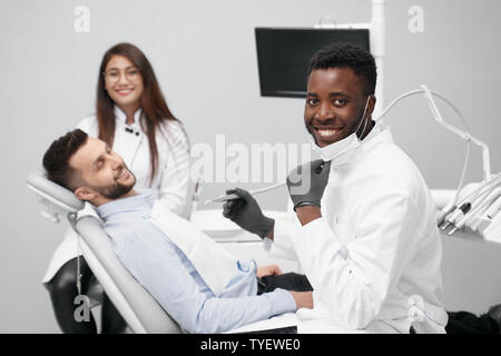 Vista laterale di esperienza africana di dentista guardando la fotocamera e sorridere mentre i denti di indurimento del paziente giovane in clinica. Medico e lavoro di assistente femminile aiutando. Concetto di stomatologia e medicina. Foto Stock