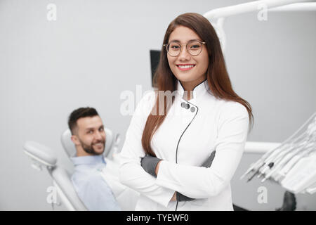 Vista frontale della bellissima femmina dentista in bianco uniforme guardando la fotocamera e in posa mentre il paziente di sesso maschile che giace sulla poltrona del dentista in background. Professional medico che lavorano nella Clinica. Foto Stock