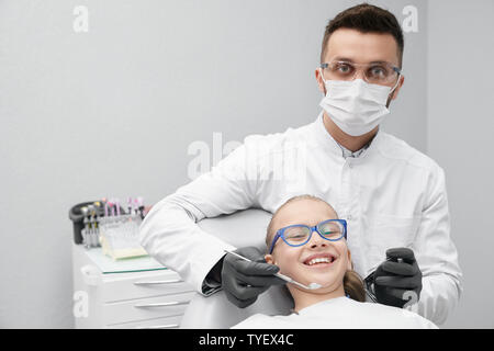 Vista frontale del giovane maschio dentista in maschera e bianco uniforme e poco paziente guardando la fotocamera e sorridente in clinica. Ragazza felice visita medico e denti di indurimento. Concetto di odontoiatria e salute. Foto Stock