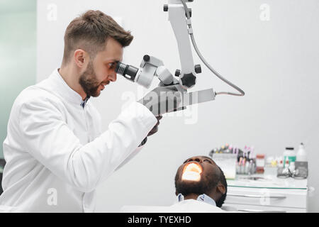 Vista dal lato del maschio professionale medico operante in clinica. Bello medico guardando le attrezzature e di esaminare i denti del paziente di sesso maschile che giace su una sedia. Concetto di stomatologia e cura. Foto Stock