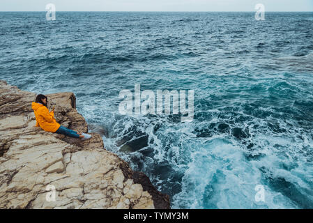 La donna posa sul bordo della scogliera alla ricerca sulle grandi onde del mare Foto Stock