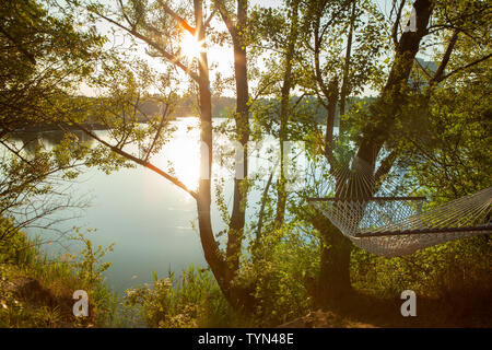 Un amaca in maglia di cotone è appeso tra due alberi che si affaccia su un lago in una luce morbida Foto Stock