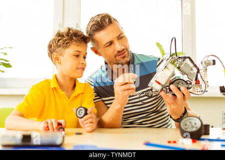 Disponibile con il tutor. Positivo uomo barbuto tenendo un robot mentre aiuta il suo allievo durante la classe Foto Stock