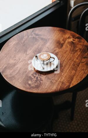 Cappuccino molto ben fatto in una tazza con arte floreale su schiuma su un tavolo rotondo in legno Foto Stock