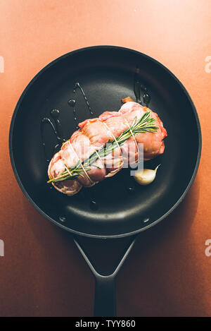 Roastbeef grezzi preparati a base di carne, caparisoned con omento di maiale grasso e rosmarino su una padella nera , vista dall'alto su un sfondo marrone Foto Stock