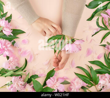 Due mani di una ragazza giovane con la pelle liscia e un mazzo di peonie rosa su uno sfondo di pesche, vista dall'alto, il concetto di moda per mano di cura della pelle, ant Foto Stock