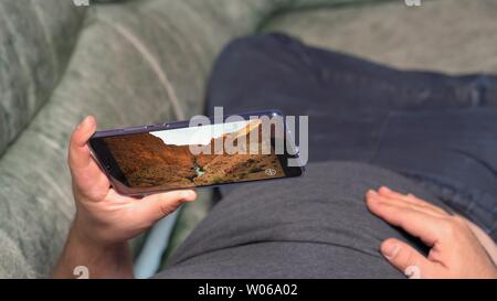 Uomo disteso sul lettino e guardare i video sui canyon valley in alta definizione tramite Huawei Mate 20 X smartphone. Il display mostra estremamente gentile e sof Foto Stock