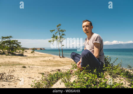 L'uomo viaggiatore rilassante sulla spiaggia di Olkhon island con vista sull'acqua del lago e delle montagne e il cielo blu. La calma e la libertà di un beauti Foto Stock