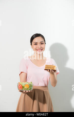 A mezza lunghezza ritratto di donna molto bella azienda piccola torta, verdure fresche. Giovani casalinga scelta di dolci o di mangiare sano - torta e insalata. Foto Stock