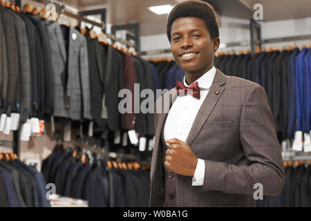 Sorridente giovane africano in posa nella boutique in camicia bianca con red bow tie, grigio tuta, guardando la fotocamera. Un sacco di costumi su appendiabiti in fila sullo sfondo. Eleganti abiti costosi per gli uomini. Foto Stock