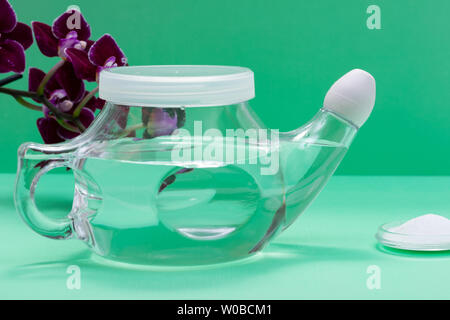 Neti Pot con Soft Comfort punta, pila di soluzione salina e viola i fiori di orchidea su sfondo verde. Lavaggio del seno. Irrigazione nasale. Foto Stock