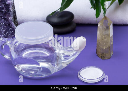 Spa sfondo viola con Neti Pot, pila di soluzione salina, arrotolato Asciugamani Bianchi, impilati pietre di basalto e foglie di bambù. Lavaggio del seno. Irrigazione nasale. Foto Stock