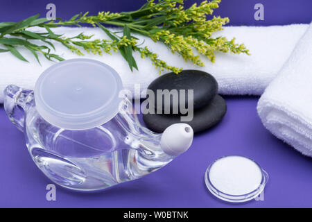 Spa sfondo viola con Neti Pot, pila di soluzione salina, arrotolato asciugamani bianchi e impilati pietre di basalto. Lavaggio del seno. Irrigazione nasale. Foto Stock