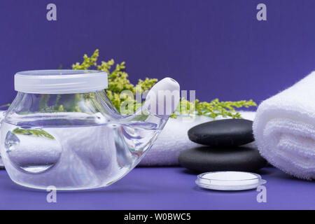 Spa sfondo viola con Neti Pot, pila di soluzione salina, arrotolato asciugamani bianchi e impilati pietre di basalto. Lavaggio del seno. Irrigazione nasale. Foto Stock