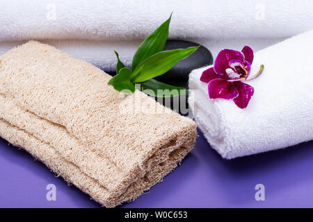 Spa Wellness Concept. Loofah naturale spugna, arrotolato Asciugamani Bianchi, impilati pietre di basalto, bambù e fiori di orchidea su sfondo viola. Foto Stock