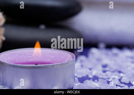 Spa Wellness Concept. Loofah naturale spugna, impilati pietre di basalto, bruciando il tè di lavanda candela luce e cosparsa di Sale di Epsom su sfondo viola. Foto Stock