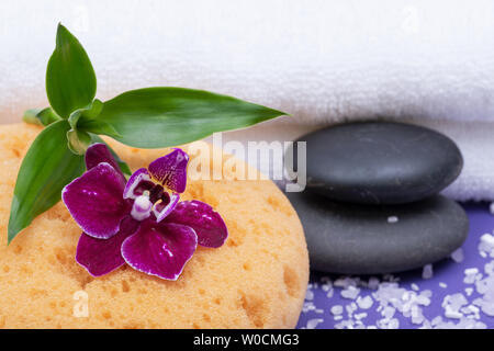 Spa Wellness Concept. Schiuma naturale Bagno & Doccia mare spugna, impilati pietre di basalto, bambù, orchidee e fiori di lavanda Sale di Epsom su sfondo viola Foto Stock