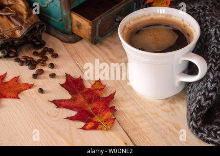 Un bianco tazza di caffè su una tavola di legno colorato con caduta foglie e chicchi di caffè con un maglione grigio e blu di una rettificatrice di antiquariato in background. Foto Stock