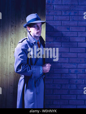 Uomo in piedi al di fuori di notte indossando un cappello e trench cercando di risolvere un mistero Foto Stock