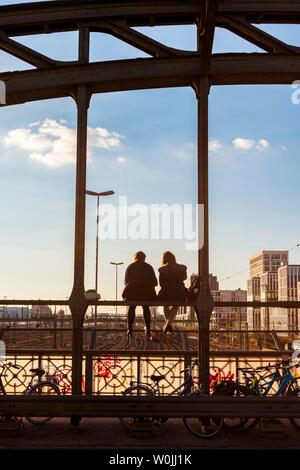 Due giovani donne seduto sulla balaustra del Haccurbrucke ponte sopra i binari ferroviari guardando in lontananza, luce posteriore, Monaco di Baviera, superiore Foto Stock